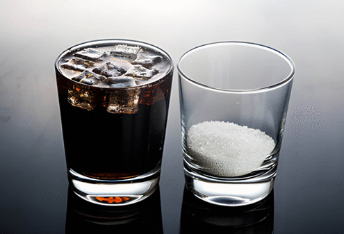 Dua gelas bening berdampingan di atas permukaan reflektif; gelas kiri berisi minuman bersoda berwarna gelap dengan es batu, gelas kanan berisi gula pasir putih dalam jumlah besar—menyoroti kandungan gula tinggi dalam satu porsi soda.