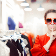 Seorang individu mengenakan pakaian merah mencolok dan kacamata besar berdiri di toko pakaian yang terang, melakukan gestur "diam" dengan jari di bibir, dikelilingi rak baju dan manekin.