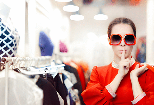 Seorang individu mengenakan pakaian merah mencolok dan kacamata besar berdiri di toko pakaian yang terang, melakukan gestur "diam" dengan jari di bibir, dikelilingi rak baju dan manekin.